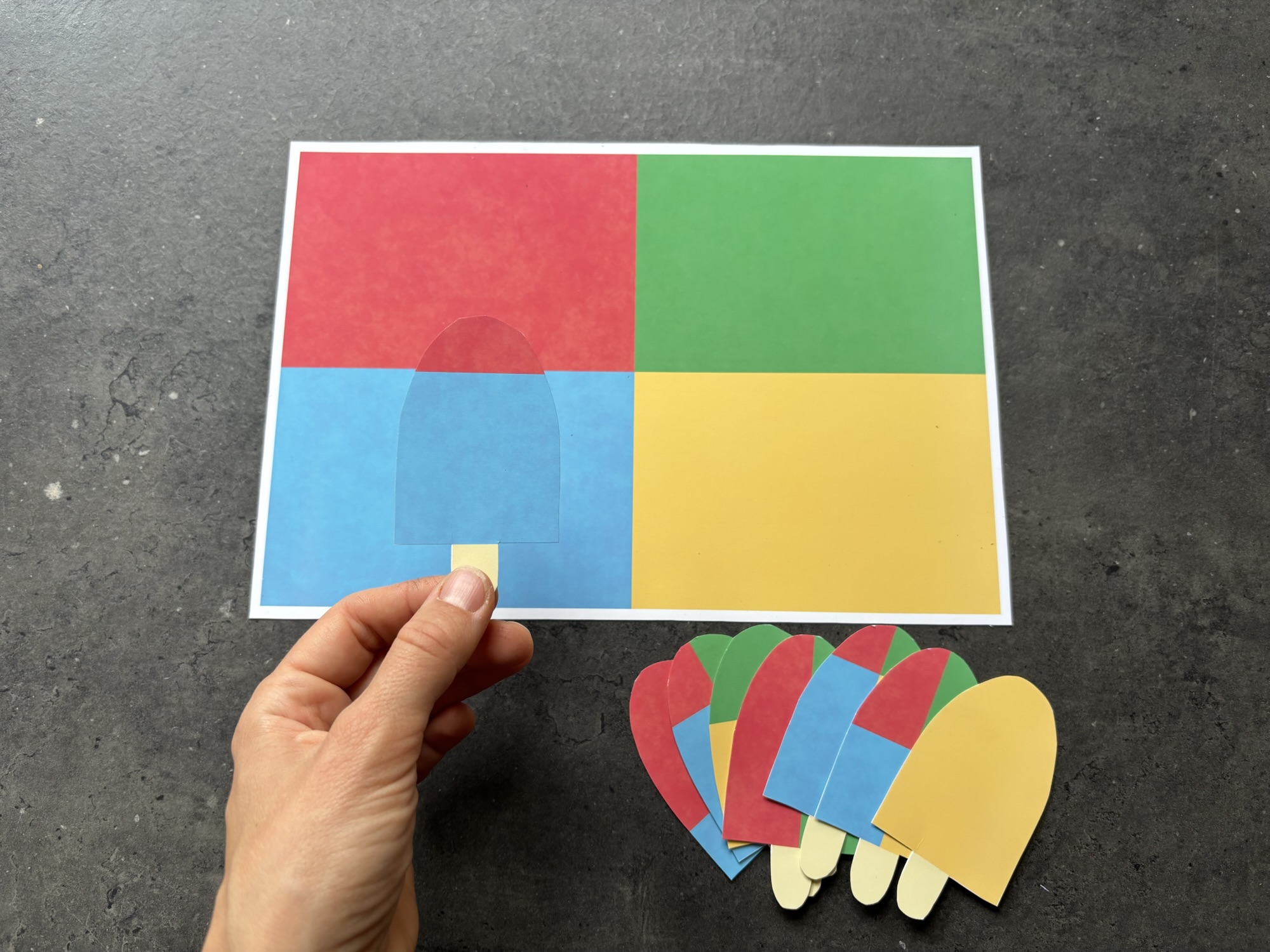 Hands-on summer popsicle matching activity for preschoolers: a hand holds a patterned popsicle cutout over a four-color master mat to practice visual discrimination and spatial logic.
